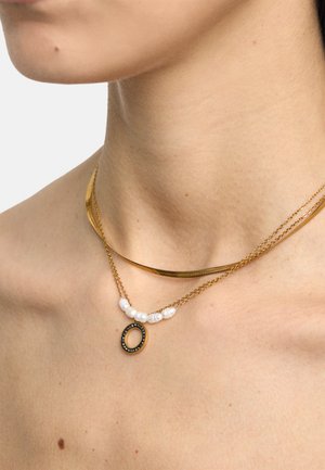 Dwa warstwowe złote naszyjniki na szyi; jeden z płaskim łańcuszkiem, drugi z białymi perłami i okrągłym złotym wisiorkiem z małymi czarnymi kamieniami.