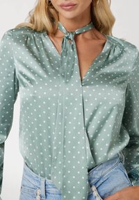 Šviesiai žalia satino bliuska su baltais taškeliais, turinti V formos iškirptę ir raištelio detalę prie iškirptės. Ilgos rankovės, laisvas stilius.