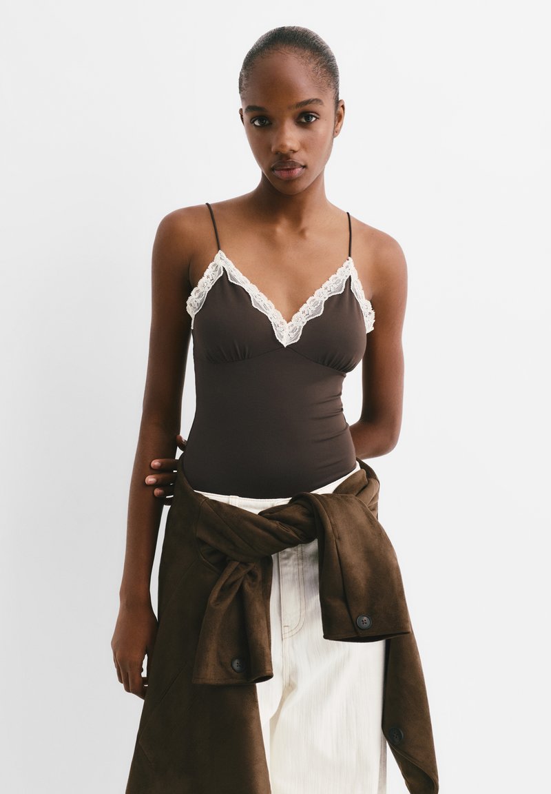 Brauner Body mit Spitzenbesatz, dünnen Trägern und anliegendem Design. Kombiniert mit einem gebundenen braunen Stoffwickel und weißen Hosen.