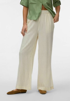 Donna che indossa pantaloni ampi color crema con sottili righe verticali nere, abbinati a scarpe marroni piatte e una camicia verde abbottonata.