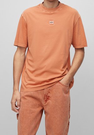T-shirt di cotone arancione con colletto tondo, maniche corte e una piccola toppa logo sul petto. Abbinato a jeans in denim arancioni coordinati.