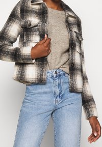 Giacca di flanella a quadri nera, bianca e grigia con due tasche sul petto. Indossata sopra un maglione beige in maglia e jeans a vita alta azzurri chiaro.