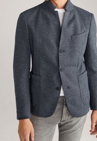 Navy textured blazer with a stand collar, single button closure, two front pockets, and a left chest pocket, paired with gray jeans.