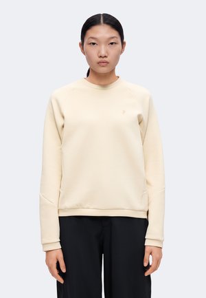 Jeune femme aux cheveux foncés attachés, portant un sweat-shirt beige à manches longues et un pantalon noir sur un fond clair uni.