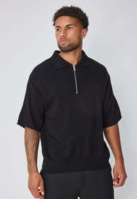 Polo en maille noire à manches courtes, col zippé et ourlet côtelé. La texture du tissu semble légèrement texturée et légère.