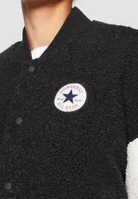 Svart fleecetröja med ett runt vitt märke som visar "Converse All Star" och en blå stjärna. Har knappstängning och kontrasterande vita ärmar.