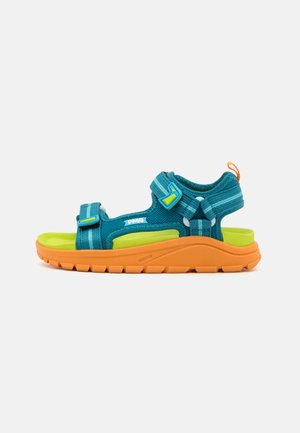 Sandalia de tela verde azulado con correas ajustables, una plantilla acolchada verde y una suela de goma naranja brillante con un patrón de relieve.