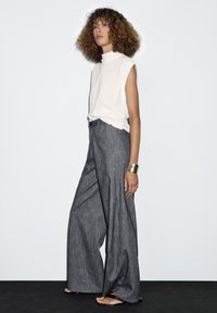 Personne avec des cheveux bouclés portant un haut sans manches blanc, un pantalon large gris, un bracelet en or et des sandales noires, se tenant contre un fond blanc.