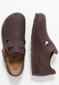 Brown leather clogs with a buckle strap, open back, and textured rubber sole. Features a rounded toe and a cushioned footbed.