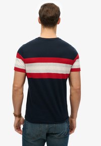 Man wearing navy blue t-shirt with red and white horizontal stripes, standing facing away, wearing blue jeans and a brown wristwatch.