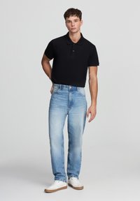 Giovane uomo che indossa una polo nera, jeans azzurri chiari e sneakers bianche, in piedi con una mano dietro la schiena su uno sfondo neutro.