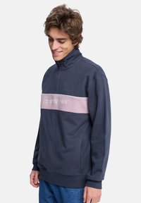 Marineblauer Fleece-Pullover mit hohem Kragen, der einen rosa horizontalen Streifen über die Brust und ein gesticktes Logo aufweist. Dezente Textur, lässige Passform.