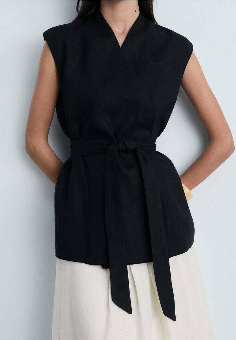 Top in lino nero con design avvolgente, maniche corte e vita con fiocco. Presenta una tasca discreta e una vestibilità rilassata, abbinato a una gonna chiara.