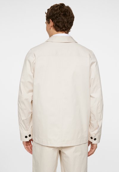 Lichtbeige jacket met een relaxte pasvorm, voorzien van een puntkraag, lange mouwen en zwarte knoopaccenten op de manchetten. Glad stoftextuur.
