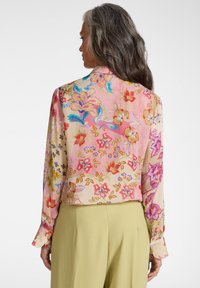 Kvinde med langt gråt hår iført en farverig blomstermønstret bluse og lysegrønne bukser, vendt væk mod en ensfarvet baggrund.