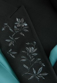 Schwarzer Stoff mit silberner floralem Stickerei an der Seite, kombiniert mit einem türkisfarbenen Stoff auf der rechten Seite. Glatte Textur, maßgeschneidertes Design.