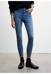 Jeans de mezclilla azul ajustados con una cintura de media altura, que cuentan con bolsillos frontales, cremallera y un desvanecimiento mínimo. Combinados con mocasines oscuros y calcetines marrones acanalados.