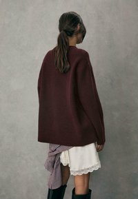 Bordeaux oversized sweater med en blød tekstur, båret over en hvid kjole med blonder. Stribet stof bundet om taljen. Sorte knæhøje støvler.