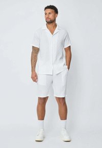 Chemise blanche à manches courtes et shorts assortis, fabriqués dans un tissu texturé avec un motif à rayures subtil, accompagnés de baskets blanches et de chaussettes.