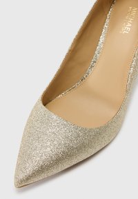 Chaussure à talon haut pointue avec finition pailletée dorée et intérieur en cuir beige portant l’étiquette Michael Kors.