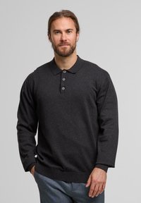 Mann mit Bart und zurückgebundenem Haar, der einen dunklen Longsleeve-Polo-Pullover und graue Hosen trägt und vor einem schlichten hellen Hintergrund steht.