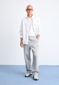 Tenue décontractée comprenant une chemise blanche à rayures, un t-shirt blanc, un pantalon de survêtement gris et des baskets légères avec des accents noirs.