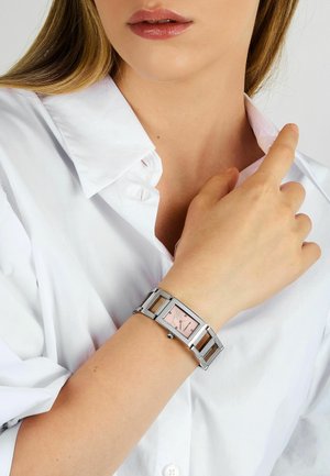 Orologio in metallo argento con quadrante rettangolare rosa, caratterizzato da dettagli ornamentali. Indossato al polso, abbinato a una camicia bianca con colletto.