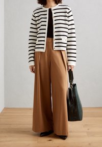 Cardigan a righe in crema e nero, abbinato a pantaloni beige a gamba larga. Porta una borsa texturizzata di un verde scuro. Tessuto morbido e vestibilità sartoriale.