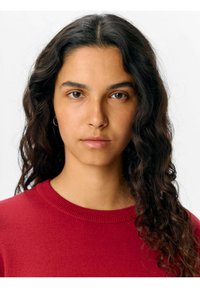 Röd tröja med ribbat rundhals. Den föreslagna modellen har lockigt mörkt hår och en naturlig sminklook, vänd mot kameran direkt.