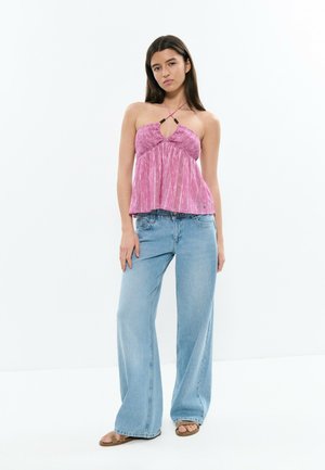 Jeune femme portant un haut rose à nouer derrière le cou et un jean large bleu clair, debout devant un fond blanc uni.