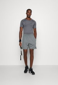 Camisa atlética ajustada gris con el logo negro de Nike, combinada con pantalones cortos gris claro y zapatillas deportivas negras. Sosteniendo una cuerda para saltar.