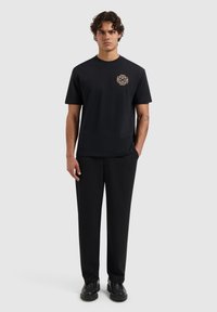 Zwarte T-shirt met een gouden intricaat logo op de linkerborst, gecombineerd met zwarte broek en zwarte schoenen. Gladde stof, relaxte pasvorm, minimalistisch ontwerp.