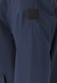 Marinblå jacka med en slät yta, prydd med en svart fyrkantig logotypapplikation på övre vänstra sidan. Enkel design, lättviktig tyg.