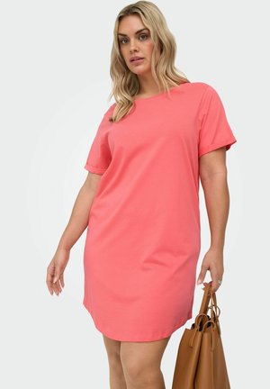 Woman with blonde hair wearing a coral short-sleeve dress, holding a brown leather handbag against a neutral background.