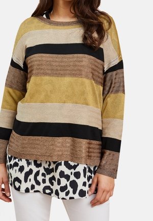 Kobieta ubrana w sweter w paski z długim rękawem w kolorach brązowym, beżowym, czarnym i musztardowym, nałożony na czarno-białą koszulę w panterkę, z białymi spodniami.