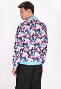 Lichtgewicht bomberjack met een levendig patroon van roze flamingo's en tropische bladeren op een donkere achtergrond. Ribgebreide manchetten met blauwe accenten.