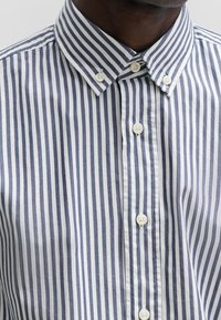 Skjorta med knäppning framtill och smala blå och vita ränder, med klassisk krage, vita knappar och en slät bomullstextur.