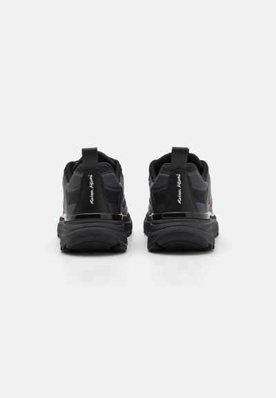 Chaussures de sport noires en matériau synthétique texturé, avec un col rembourré et une languette au niveau du talon. Semelle épaisse avec des motifs géométriques subtils.