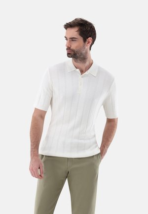 Polo a maniche corte realizzato in tessuto bianco a coste, con colletto piatto e una chiusura a due bottoni, indossato con pantaloni verde oliva.