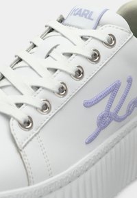 Λευκό αθλητικό παπούτσι με ασημένιες τρύπες, διαθέτει λευκές κορδόνια και υφή στη σόλα. Σημαντικό στοιχείο είναι το ανοιχτό μωβ κεντητό λογότυπο "K" στο πλάι.