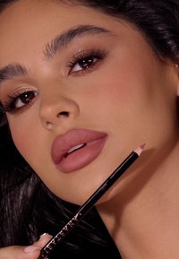 Konturówka do ust w stonowanym różowym odcieniu trzymana blisko zbliżenia twarzy, z pełnymi ustami i delikatnie rozświetloną skórą; czarne i srebrne opakowanie.