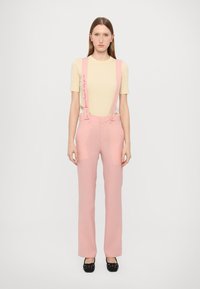Pink trousers with suspenders featuring text accents, paired with a beige t-shirt. Fabric has a smooth texture and tailored shape. Black shoes complete the look.