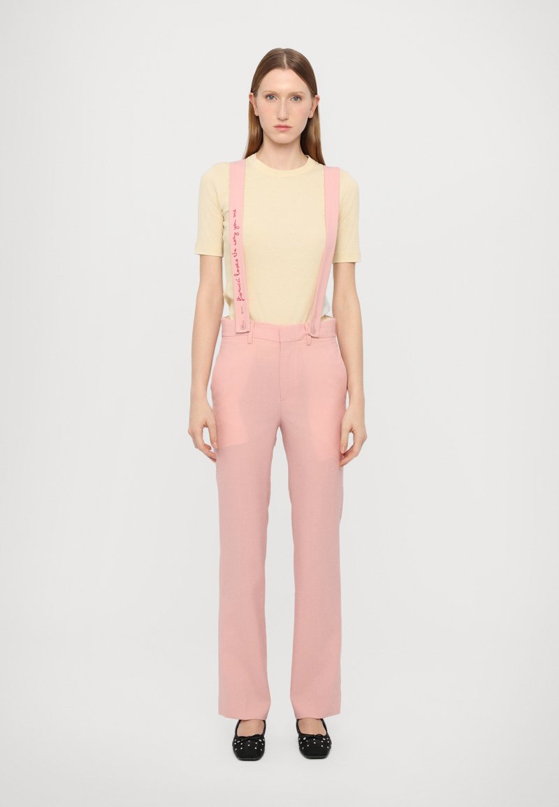 Pink trousers with suspenders featuring text accents, paired with a beige t-shirt. Fabric has a smooth texture and tailored shape. Black shoes complete the look.