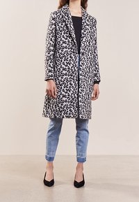 Lapjesjasje met panterprint in een gestructureerde stof, met een notch-kraag, knoopsluiting en lange mouwen, gecombineerd met lichtblauwe jeans en zwarte hakken.