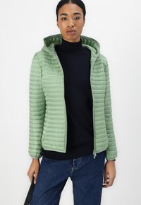 Lysegrøn pufferjakke med lynlås i front og hætte, med vandret quiltning og grå accenter. Kombinationen er med en navy turtleneck.
