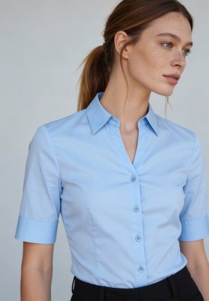 Junge Frau mit Sommersprossen, die ein hellblaues Kurzarm-Hemden und schwarze Hosen trägt, schaut nach rechts vor einem schlichten Hintergrund.