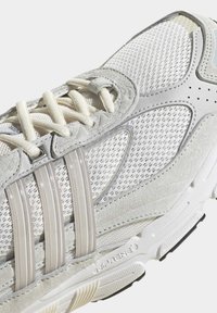 Fehér sportcipő hálós felsőrésszel és szürke velúr díszítésekkel. Három átlátszó, vízszintes csíkkal, valamint párnázott talppal rendelkezik, amelyen „adiPRENE” márkajelzés található.