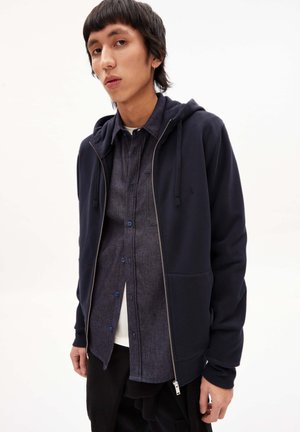 Marineblauwe zip-up hoodie met verstelbare koord, gecombineerd met een donkerblauw overhemd met knopen. Heeft een slank ontwerp en een soepele stofstructuur.