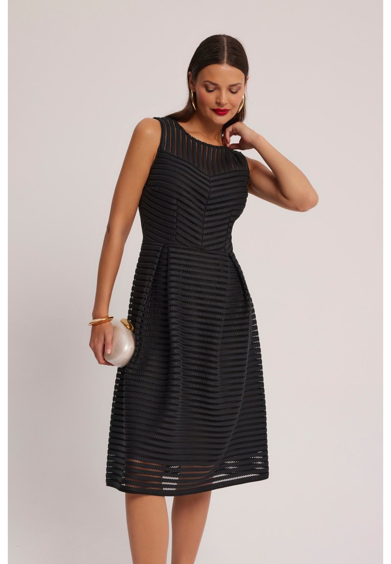 Czarna sukienka z dopasowanym gorsetem, przezroczystą paskowaną teksturą i rozkloszowanym dołem. Gładki materiał o subtelnym połysku; zestawiona z okrągłą kopertówką.