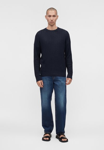 Άνδρας με κοντά μαλλιά, ντυμένος με ναυτικό μπλε πουλόβερ πλεκτό με σχέδιο κόμπων, μπλε τζιν και μαύρα σανδάλια, στέκεται μπροστά σε απλό φωτεινό φόντο.